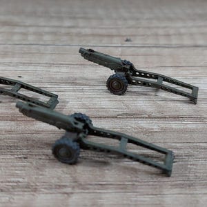Peut inclure: Trois canons miniatures vert olive avec des roues noires sont disposés sur une surface en bois. Les canons ont un design détaillé, comprenant le canon, l'affût et les roues. L'arrière-plan est une texture de bois brun clair.