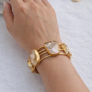 May include: Gold-toned bracelet featuring four irregularly shaped, clear, and iridescent stones. The bracelet has a multi-strand design and is worn on a wrist. A cup of coffee, a white saucer, and a rose are in the background.