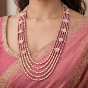 May include: A multi-strand necklace featuring alternating strands of pink beads and white pearls. The necklace is adorned with gold-colored floral accents and is worn over a pink garment.
