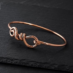 Puede incluir: Un brazalete rígido de tono oro rosa con un acabado liso y pulido. La pulsera está diseñada con un alambre continuo que forma un detalle decorativo en forma de nudo en espiral en el centro, creando una estructura circular de bucle abierto.