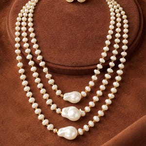 May include: A three-strand pearl necklace set with matching stud earrings. The necklaces feature small, round pearls with larger, baroque-shaped pearls at the center. The jewelry is set against a brown background.