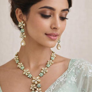 Puede incluir: Elegante conjunto de joyas florales con collar y pendientes. Las piezas doradas están adornadas con piedras preciosas transparentes y verde claro, creando un aspecto delicado y sofisticado. Los pendientes tienen forma de lágrima.