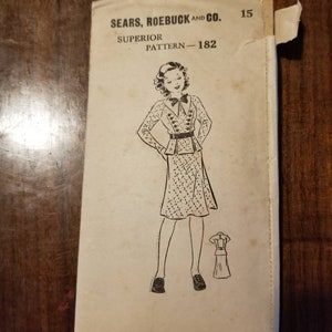 Op de afbeelding: Vintage naaipatroon van Sears, Roebuck and Co. voor een jurk. Het patroonnummer is 182. Het patroon toont een vrouw die een jurk draagt met een getailleerd lijfje en een volle rok. De jurk heeft een riem en knopen aan de voorkant. Het patroon bevat ook een kleine illustratie van de achterkant van de jurk.