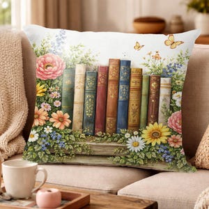 May include: Decorative throw pillow featuring a watercolor design of antique books, flowers, and butterflies. The pillow has a white background with colorful illustrations of a floral garden and a row of vintage books.