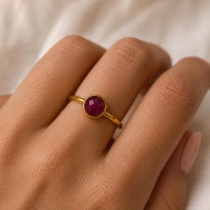Könnte beinhalten: Ein goldener Ring mit einem einzelnen, runden, facettierten tiefrosa Edelstein in einer glatten Zargenfassung. Die Ringschiene ist dünn und poliert und zeigt ein schlichtes Design, das die leuchtende Farbe und den geometrischen Schliff betont.