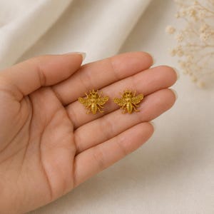 May include: A pair of gold-toned bee-shaped stud earrings. Each earring features a detailed, textured body with wings extended outward, showcasing a metallic finish and a small crown-like detail on the head of the bee, designed for earlobe wear.