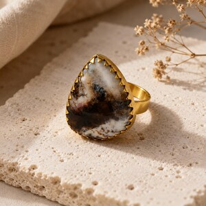 Könnte beinhalten: Ein goldfarbener Ring mit einem großen, birnenförmigen Stein, der ein marmoriertes Muster in Weiß, Dunkelbraun und Schwarz aufweist. Der Stein ist in einer gezackten Metallfassung eingefasst und die Ringschiene besteht aus glattem Metall.