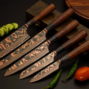 Puede incluir: Un juego de cinco cuchillos de cocina con mangos de madera oscura y hojas de acero de Damasco color cobre con patrones ondulados. Los cuchillos están dispuestos en diagonal junto a pepinos en rodajas, chiles verdes y un tomate.