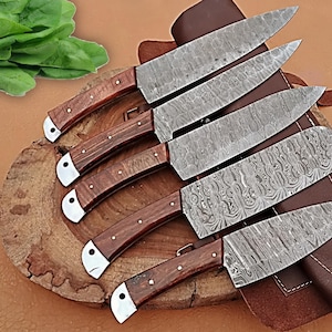 May include: A set of six Damascus steel kitchen knives with wooden handles and silver bolsters. The blades have a unique patterned design. The knives are arranged on a wooden cutting board with a leather sheath.