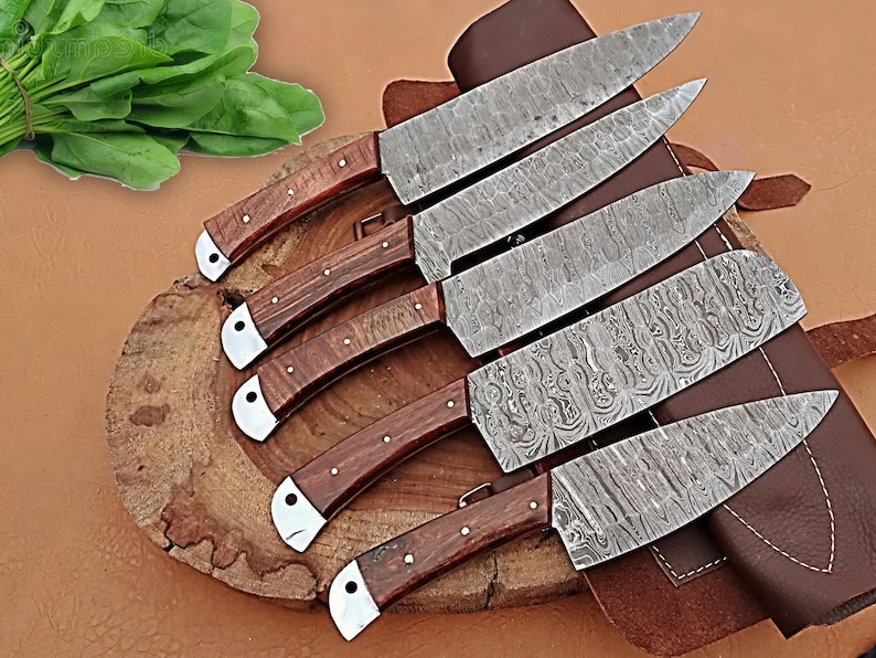 May include: A set of six Damascus steel kitchen knives with wooden handles and silver bolsters. The blades have a unique patterned design. The knives are arranged on a wooden cutting board with a leather sheath.
