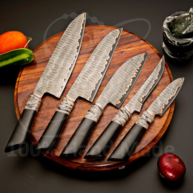 May include: A set of five handcrafted kitchen knives with textured blades and black handles. The blades have a wavy edge with a copper-colored accent. The knives are arranged on a wooden cutting board.
