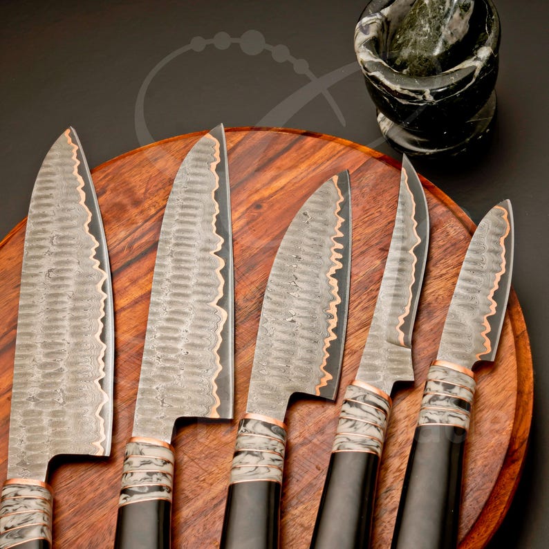 May include: Six handcrafted kitchen knives with textured blades and copper accents are arranged on a wooden cutting board. The knives have black handles with decorative white and gray bands. A mortar and pestle are in the background.