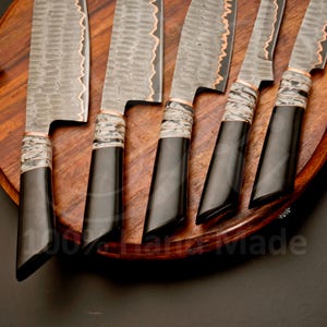 May include: Set of six handcrafted kitchen knives with textured steel blades and black handles. The blades have a wavy edge with copper accents. The handles are black with a white and gray pattern near the blade. The knives are arranged on a wooden surface.