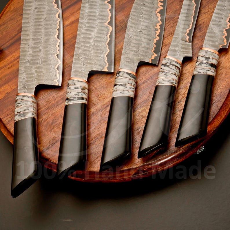 May include: Set of six handcrafted kitchen knives with textured steel blades and black handles. The blades have a wavy edge with copper accents. The handles are black with a white and gray pattern near the blade. The knives are arranged on a wooden surface.