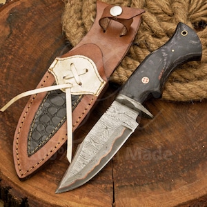 May include: A Damascus steel knife with a black handle and a brown leather sheath. The blade has a unique pattern, and the sheath features a decorative stitched design with a black, textured accent. The knife is designed for outdoor use.