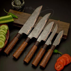 Op de afbeelding: Een set van vijf handgemaakte keukenmessen met donkere houten handvatten en stalen messen met patronen. De messen hebben een koperkleurige rand. Gesneden komkommers en tomaten zijn zichtbaar.