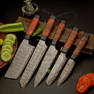 May include: Set of six Damascus steel kitchen knives with wooden handles. The blades have a unique pattern, and the handles are a reddish-brown color. The knives are arranged on a wooden board, with sliced cucumbers and tomatoes nearby.