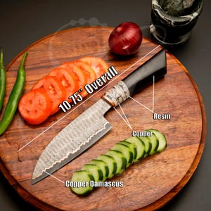 May include: A 10.75" overall Damascus steel chef's knife with a black resin handle and copper accents. The knife is displayed on a wooden cutting board with sliced tomatoes, cucumbers, and an onion. The blade has a copper damascus design.