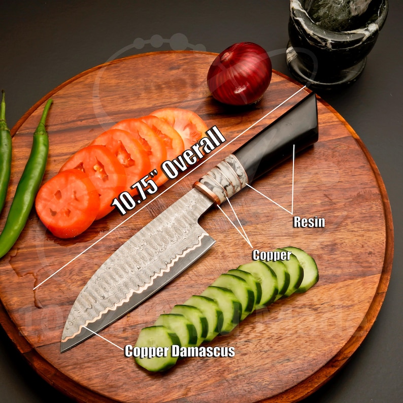 May include: A 10.75" overall Damascus steel chef's knife with a black resin handle and copper accents. The knife is displayed on a wooden cutting board with sliced tomatoes, cucumbers, and an onion. The blade has a copper damascus design.