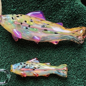 May include: Two glass fish sculptures with iridescent colors, including yellow, pink, and blue. The fish have detailed fins and spots, and are displayed on a green surface. One fish is larger than the other.