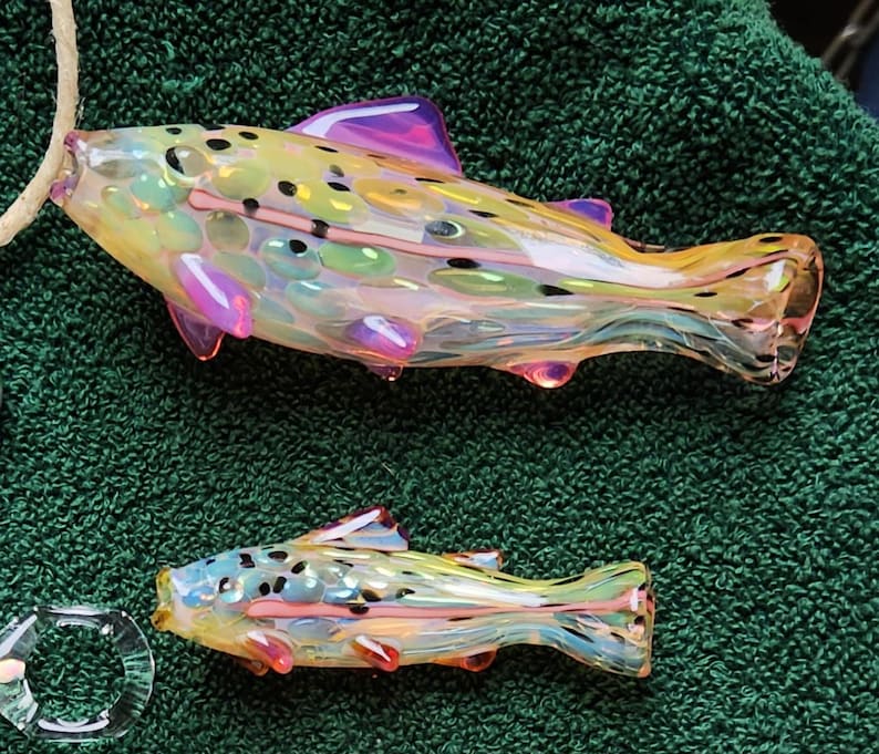 May include: Two glass fish sculptures with iridescent colors, including yellow, pink, and blue. The fish have detailed fins and spots, and are displayed on a green surface. One fish is larger than the other.