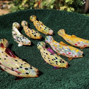 May include: A collection of seven colorful, glass fish figurines. Each fish has a unique pattern of spots and colors, including yellow, orange, red, and black. The fish are arranged on a green surface, with sunlight illuminating them.