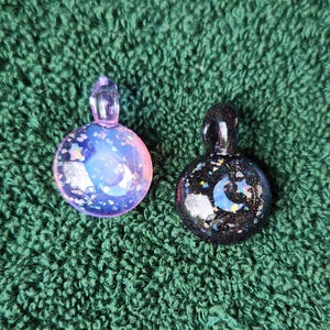 May include: Two glass pendants with celestial designs. One is a translucent pink and purple with a moon and stars, the other is black with a blue moon and stars. Both have a loop at the top for a chain.