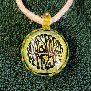 May include: A round pendant with the words "ADESPREA RITZI" in black script on a yellow background. The pendant has a green and yellow border and is suspended from a white cord. The pendant is made of glass.