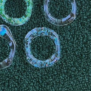 May include: Close-up of several clear resin rings with embedded glitter. The rings have a circular shape with a flat top and are filled with blue and green glitter. The background is a dark green fabric.