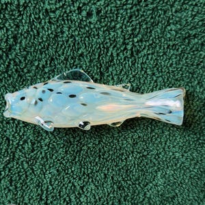 May include: A glass fish figurine with a light blue body and black spots. The fish has clear fins and a tail, and is resting on a green textured surface. The figurine is approximately 6 inches long.