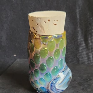 Op de afbeelding: Een kleurrijke glazen pot met een kurken stopper. De pot is versierd met groene en blauwe ovale vormen. De bodem van de pot heeft een wervelend blauw ontwerp. De kurk is lichtbruin.