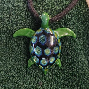 May include: A handcrafted glass turtle pendant with a black cord. The turtle has a green head, flippers, and tail. The shell is a mix of black, blue, and green iridescent colors. The pendant is a unique piece of jewelry.