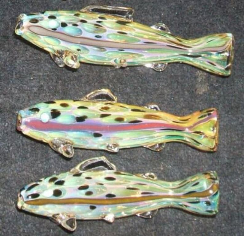 May include: Three glass fish figurines. Each fish is a different color, with a mix of green, yellow, and blue. The fish have black spots and clear fins. The fish are arranged in a row.