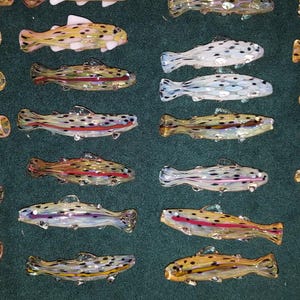 May include: An assortment of colorful glass fish figurines. The fish vary in color, including yellow, brown, and white, with black and red accents. The fish are arranged on a dark green surface.