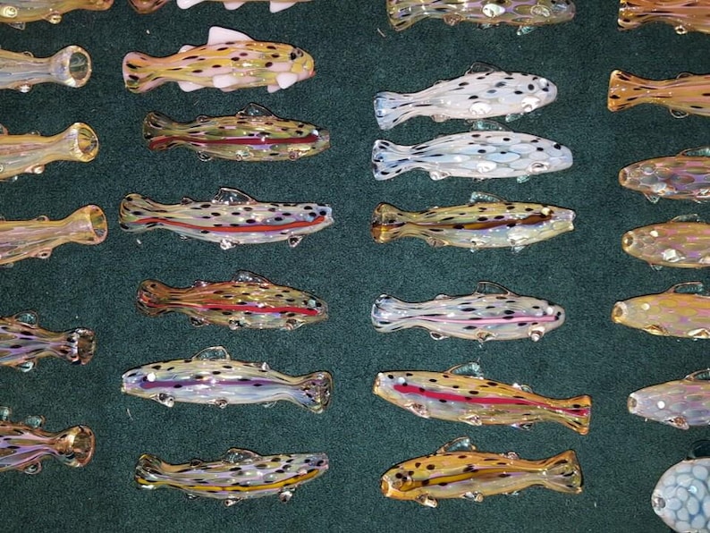 May include: An assortment of colorful glass fish figurines. The fish vary in color, including yellow, brown, and white, with black and red accents. The fish are arranged on a dark green surface.