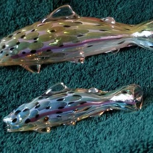 May include: Two iridescent glass fish figurines. The fish have a speckled pattern in shades of green, blue, and purple. The fins and tails are clearly defined, and the overall design is detailed. The fish are displayed on a teal green surface.