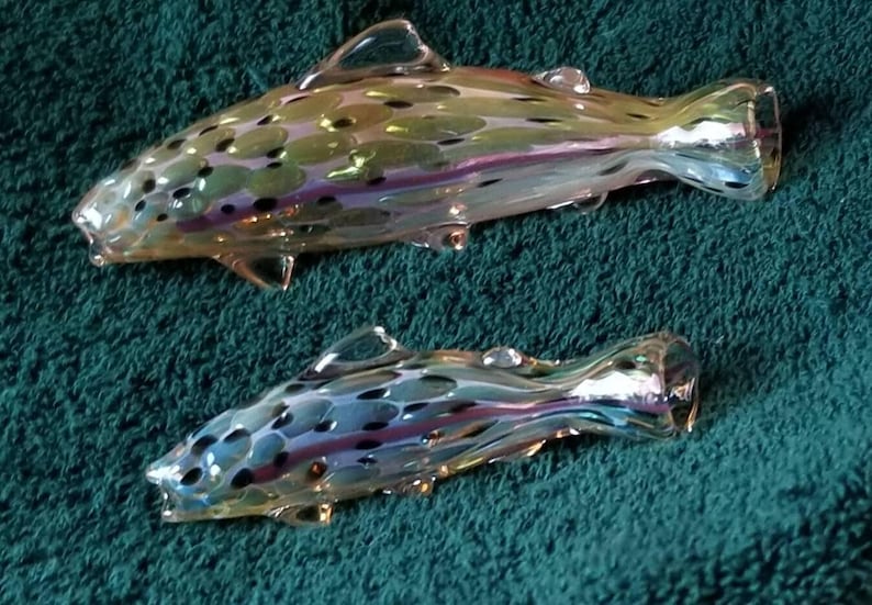May include: Two iridescent glass fish figurines. The fish have a speckled pattern in shades of green, blue, and purple. The fins and tails are clearly defined, and the overall design is detailed. The fish are displayed on a teal green surface.