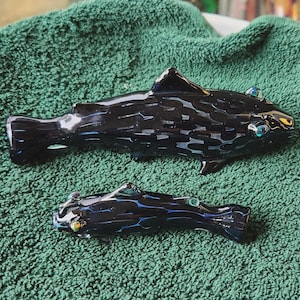 May include: Two black glass fish sculptures with blue and green accents. The larger fish has a dorsal fin and a tail fin. The smaller fish has a similar design. Both are placed on a green textured surface.