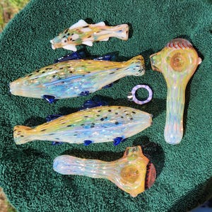 May include: A collection of colorful glass art pieces, including fish-shaped pipes and a ring. The fish pipes have yellow, blue, and green hues with dark blue fins. The ring is a light purple color. The pieces are arranged on a green towel.