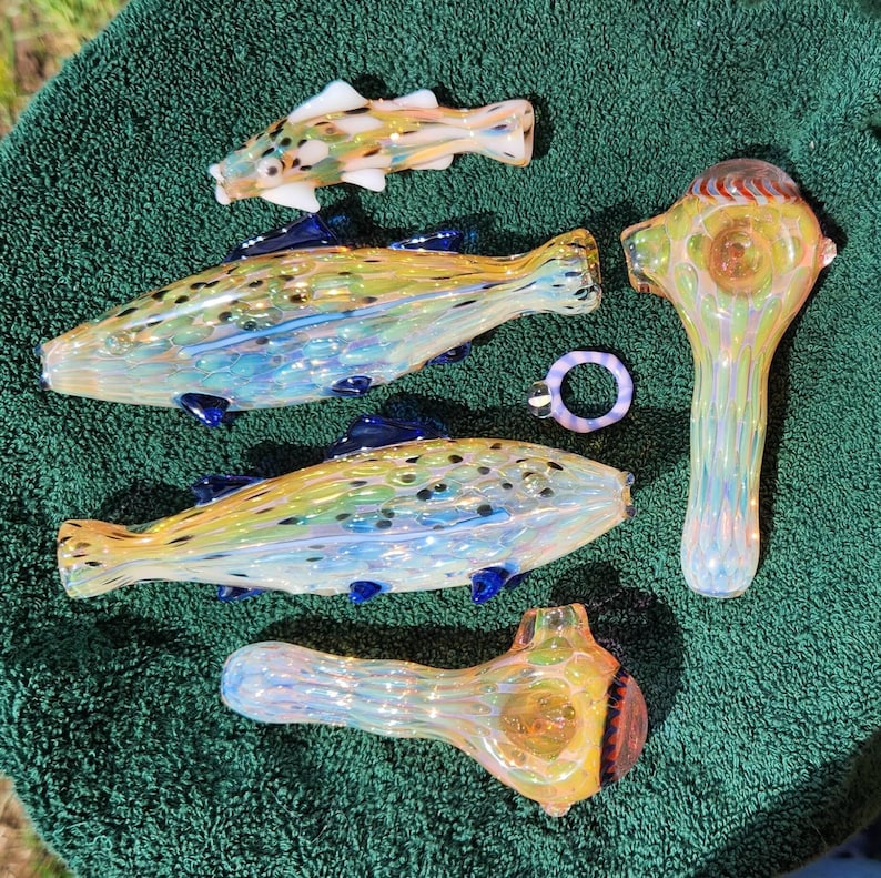 May include: A collection of colorful glass art pieces, including fish-shaped pipes and a ring. The fish pipes have yellow, blue, and green hues with dark blue fins. The ring is a light purple color. The pieces are arranged on a green towel.