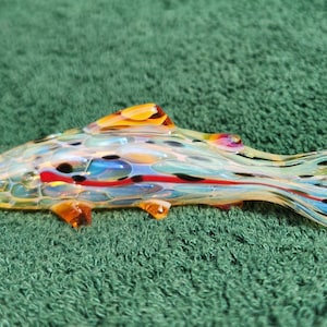 May include: A colorful glass fish sculpture with yellow, orange, red, blue, and black accents. The fish has fins and a tail, and is resting on a green surface. The glass is translucent, allowing light to pass through.
