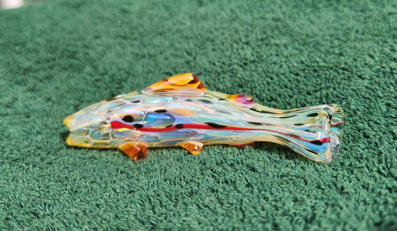 May include: A colorful glass fish sculpture with yellow, orange, red, blue, and black accents. The fish has fins and a tail, and is resting on a green surface. The glass is translucent, allowing light to pass through.