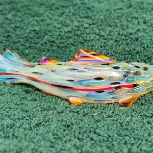 May include: A colorful glass fish-shaped pipe with a clear body, featuring blue, red, and yellow accents. The pipe has a textured surface and is set against a green background. The fish has a fin and a detailed design.