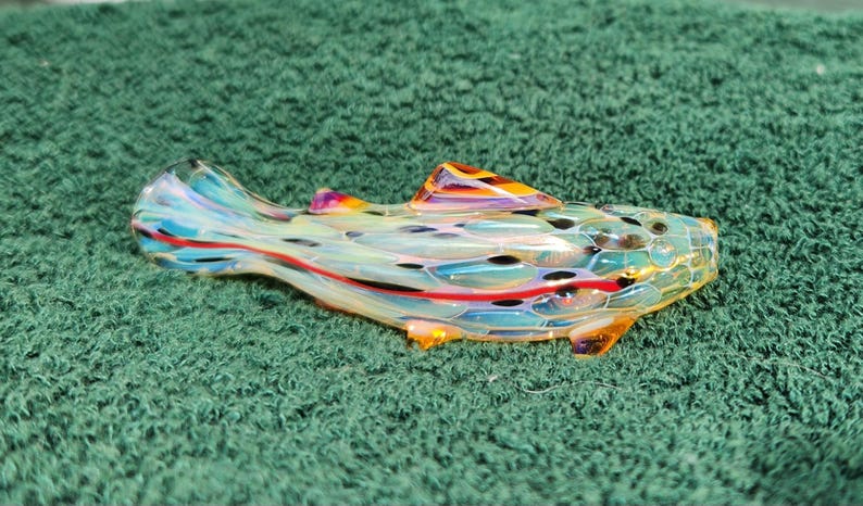 May include: A colorful glass fish-shaped pipe with a clear body, featuring blue, red, and yellow accents. The pipe has a textured surface and is set against a green background. The fish has a fin and a detailed design.
