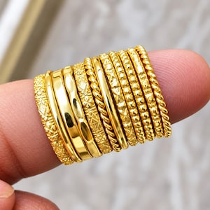 Puede incluir: Un conjunto de diez anillos de tono dorado con diversas texturas y diseños, incluyendo bandas lisas, patrones de cuerda trenzada, superficies con cuentas y grabados geométricos intrincados, dispuestos lado a lado en formación.