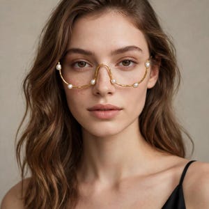Può includere: Un accessorio per occhiali in metallo dorato con un design a montatura curva senza bordi che poggia sul ponte del naso. Il sottile filo d'oro testurizzato è accentuato da sei piccole perle bianche rotonde distanziate lungo l'arco inferiore.