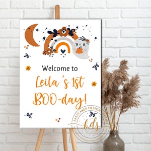 May include: Welcome to Leila's 1st BOO-day! A white sign with a cute ghost holding a pumpkin in front of a rainbow with orange, yellow, and white stripes. The sign has a black and white floral design and stars.