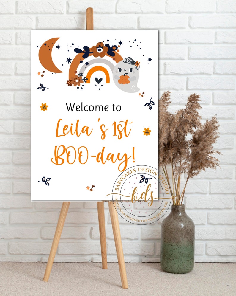 May include: Welcome to Leila's 1st BOO-day! A white sign with a cute ghost holding a pumpkin in front of a rainbow with orange, yellow, and white stripes. The sign has a black and white floral design and stars.
