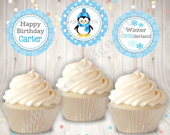 May include: Three white cupcakes with blue and white snowflake and penguin themed cupcake toppers. The toppers say "Happy Birthday Carter", "Winter ONEderland", and "Baby Cakes".