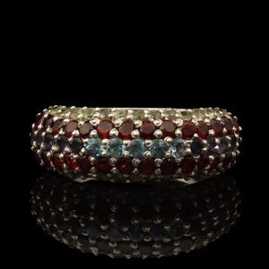 Puede incluir: Un anillo de plata con una banda de pequeñas piedras preciosas redondas. El anillo presenta filas de piedras rojas, azul claro y verdes. El anillo está sobre un fondo negro, con su reflejo visible debajo.